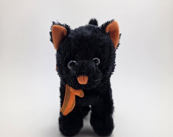 Puede incluir: Un gato negro de peluche con orejas, nariz y lazo naranja. El gato de peluche tiene ojos blancos y está de pie. Un juguete de peluche lindo y tierno.