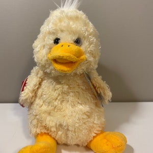 May include: A yellow plush duck with a white tuft of feathers on its head. The duck has a yellow beak and orange feet. The duck is sitting on a white surface.