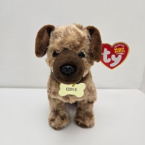 May include: Brown and tan plush dog toy with a black collar and a yellow tag that says "ODIE". The toy has a Ty Beanie Babies tag attached to it.