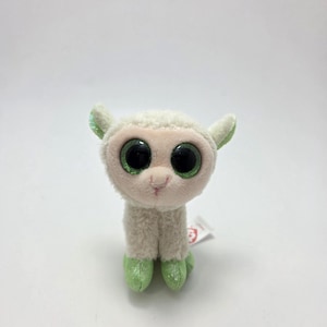May include: A plush toy llama with large, round, black eyes with green glitter accents. The llama has a cream-colored body, green glitter accents on its ears and feet, and a small pink nose. The toy is on a white background.