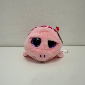 May include: Pink and purple plush turtle with large, sparkly eyes. The turtle has a small, pink nose and a pink mouth. The turtle is a Beanie Boo keychain.