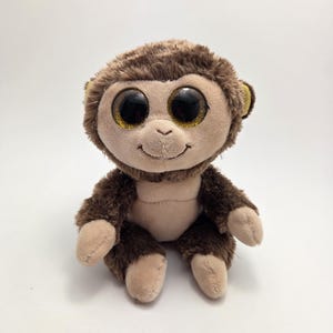 May include: A plush monkey toy with brown fur, tan face, hands, and feet. The monkey has large, round eyes with gold glitter accents and a small, smiling mouth. The toy is sitting upright on a white surface.