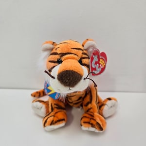 Puede incluir: Un tigre de peluche naranja y negro con la cara blanca y una etiqueta azul y roja que dice "Beanie Babies" y "2002".