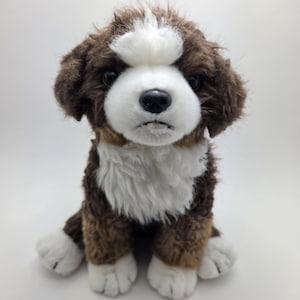 May include: A soft toy dog with brown and white fur, in a sitting position. The toy has a white face, chest, and paws, with brown fur on its head and body. The dog has black eyes and a black nose.