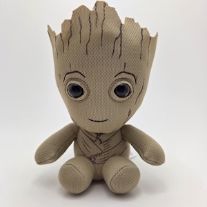 May include: A plush toy of the character Groot from Guardians of the Galaxy. The toy is tan with a textured fabric, large black eyes, and a small smile. It has a tree-like head and is in a sitting position.