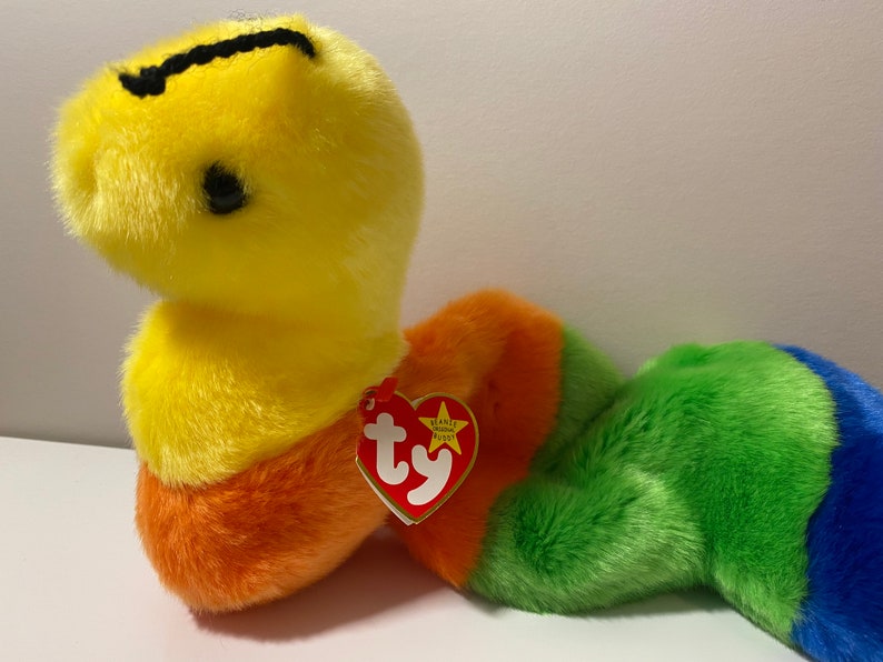Op de afbeelding: Een gele, oranje, groene en blauwe pluche slang met een zwarte streep op zijn kop. De slang heeft een rood Ty Beanie Baby-label aan zijn zijde bevestigd. Het label heeft een witte ster en het woord "Ty" in wit.