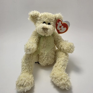 Può includere: Un orsacchiotto di peluche color crema con pelliccia arruffata, occhi scuri e naso ricamato marrone. L'orso presenta un nastro trasparente verde chiaro legato attorno al collo e un'etichetta Ty rossa e gialla a forma di cuore.