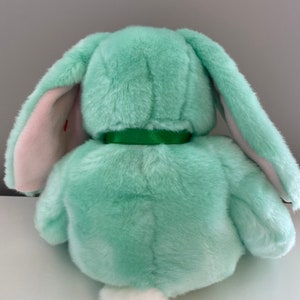 Puede incluir: Un peluche de conejo de color verde menta, visto desde atr&aacute;s. El conejo tiene largas orejas ca&iacute;das con el interior rosa, una cola blanca y esponjosa, y una cinta verde alrededor del cuello. El juguete est&aacute; hecho de un material suave y afelpado.