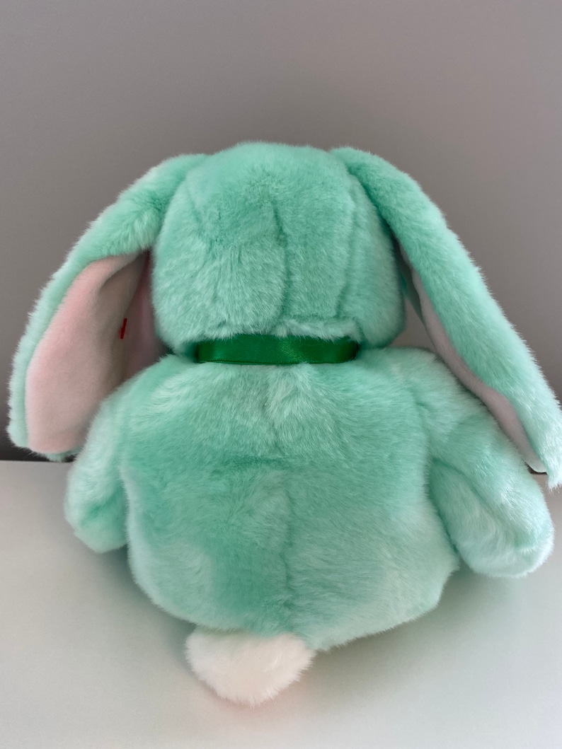 Puede incluir: Un peluche de conejo de color verde menta, visto desde atr&aacute;s. El conejo tiene largas orejas ca&iacute;das con el interior rosa, una cola blanca y esponjosa, y una cinta verde alrededor del cuello. El juguete est&aacute; hecho de un material suave y afelpado.
