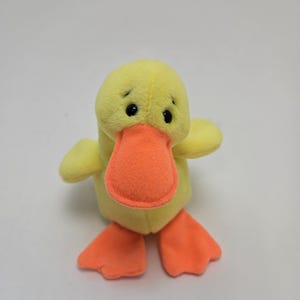 May include: A yellow plush duck with orange feet and beak. The duck has black eyes and a small, round body.