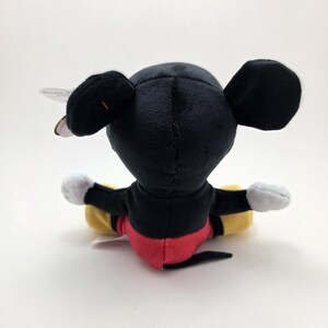 May include: A plush Mickey Mouse toy, viewed from the back, with black ears, a black head, and a red and black body. The toy has white gloves and yellow and black legs. A tag is attached to the ear.