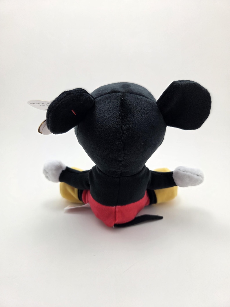 May include: A plush Mickey Mouse toy, viewed from the back, with black ears, a black head, and a red and black body. The toy has white gloves and yellow and black legs. A tag is attached to the ear.