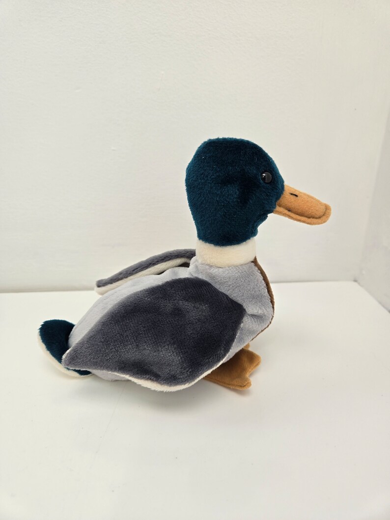 Pu&ograve; includere: Un'anatra di peluche con la testa turchese, il corpo grigio e il becco e le zampe marroni.