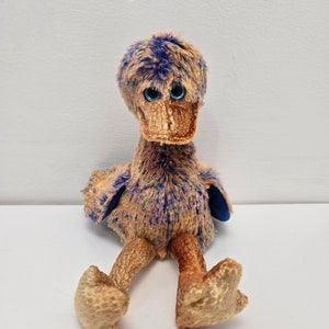 May include: A colorful plush toy duck with blue eyes and a gold beak. The duck is sitting with its legs outstretched and has a multi-colored fur pattern.