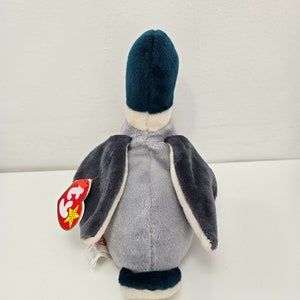 Pu&ograve; includere: Un'anatra di peluche grigia e blu con la pancia bianca e un'etichetta Ty attaccata all'ala. L'anatra &egrave; seduta su una superficie bianca.
