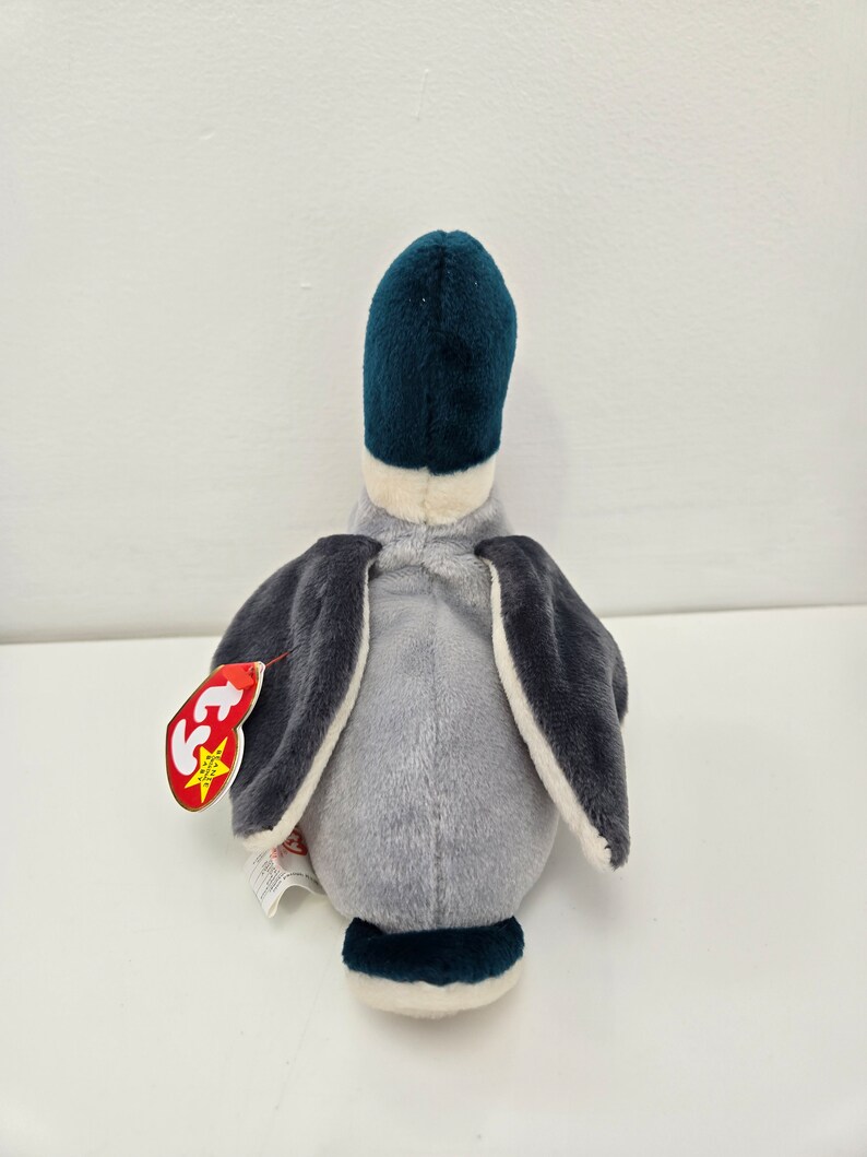 Pu&ograve; includere: Un'anatra di peluche grigia e blu con la pancia bianca e un'etichetta Ty attaccata all'ala. L'anatra &egrave; seduta su una superficie bianca.