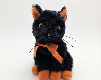 Puede incluir: Un gato de peluche negro con detalles naranjas en las orejas y las patas. Tiene un pequeño lazo naranja y bigotes. El gato está sentado erguido sobre un fondo blanco.