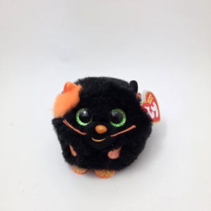 May include: A plush, round black cat toy with orange accents on its ears and paws. The cat has large, glittery green eyes, an orange nose, and orange whiskers. The toy has a red tag attached.