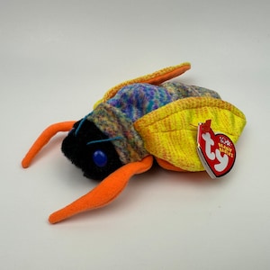 May include: A colourful plush insect toy with a black head, orange antennae, and a yellow and blue body. The toy has a red tag with white text. The toy is a Beanie Baby.
