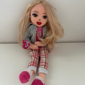 Op de afbeelding: Een pluche pop met blond haar, die een grijs jack, een roze shirt met het nummer 30, een geruite broek en roze pantoffels draagt.