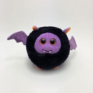 May include: A plush bat toy with a round black body and purple face, wings, and accents. The bat has large orange eyes and small orange ears. The wings are a lighter shade of purple. The toy is designed to look like a friendly Halloween character.