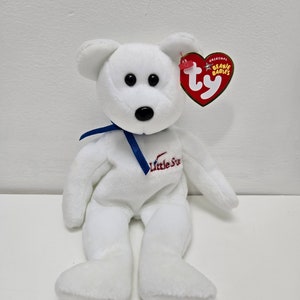 May include: White plush bear with a blue ribbon around its neck. The bear has black eyes and a black nose. The bear is sitting on a white surface. The bear has a Ty Beanie Babies tag attached to its ear. The tag is red and white with the words "Original Beanie Babies" and the Ty logo. The bear has the words "Little Star" embroidered on its chest.