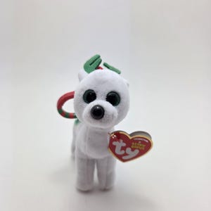 May include: A white plush reindeer toy with green antlers and a red and green loop. The toy has large, round, green eyes and a black nose. A red heart-shaped tag with the text "ORIGINAL Beanie Babies" is attached.