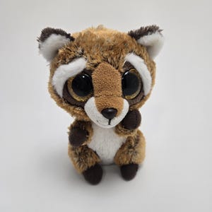 May include: A brown and white plush raccoon with large, glittery eyes. The raccoon is sitting with its paws tucked in front of it.