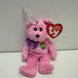 May include: Pink plush Easter bunny beanie baby with a purple ribbon and an embroidered Easter egg on its chest. The beanie baby has a Ty tag that reads "Basket Beanies Ty".