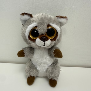 May include: A gray and white plush raccoon with large, sparkly gold eyes. The raccoon is sitting with its paws in front of it.