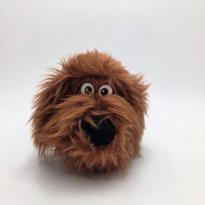 Op de afbeelding: Een pluchen speelgoedhond met ruige bruine vacht, grote ronde ogen en een zwarte neus. Het speelgoed is ontworpen om op een hondenras te lijken, met een pluizig uiterlijk.