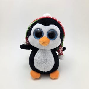 May include: A black and white penguin plush toy wearing a red and green knit hat with a white pom-pom. The penguin has large blue eyes and an orange beak.