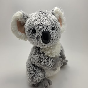 Może przedstawiać: Pluszowy miś koala z szaro-białym futrem, czarnym nosem i dużymi, okrągłymi oczami. Koala ma puszyste uszy z różowymi detalami wewnątrz i siedzi wyprostowany. Miękka, przytulna pluszowa zabawka.