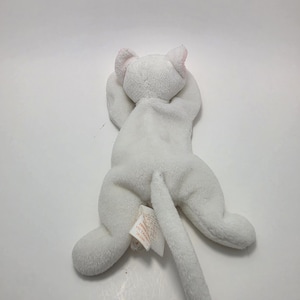 以下が含まれることがあります： 頭の上に前足を伸ばした伏せの姿勢の白い猫のぬいぐるみ。柔らかい白い生地で作られており、内側が淡いピンク色の尖った耳と長い尻尾が特徴です。後ろ側には白い布製のタグが付いています。