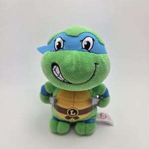 May include: A plush toy of Leonardo from Teenage Mutant Ninja Turtles. The toy is green with a blue mask, blue armbands, and a brown belt. The turtle has a smiling face and is standing upright.