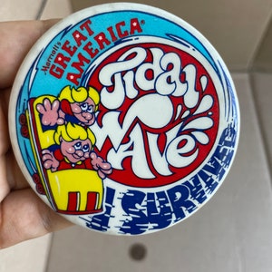 May include: A white button with a blue, red, and yellow design. It features a cartoon image of two people riding a yellow roller coaster with the words "Tidal Wave! Surf Waves" and "Great America" in a circle around the image.