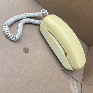 May include: A vintage yellow rotary phone with a coiled cord. The phone is sitting in a cardboard box.