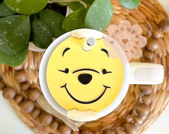 Może przedstawiać: Biały kubek podróżny z żółtą pokrywką z twarzą Winnie the Pooh. Pokrywka ma otwór na słomkę i czarny kontur wokół twarzy. Kubek ma biały uchwyt.