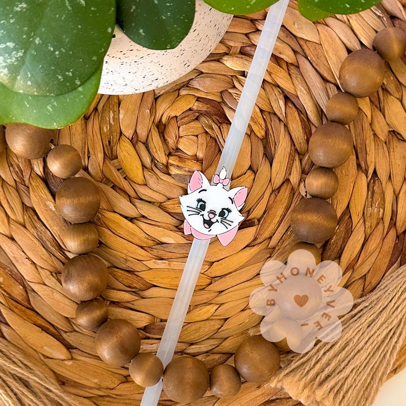 May include: A white cat with pink ears and a pink bow, with a white straw behind it. The cat is on a woven brown wicker surface with brown wooden beads.