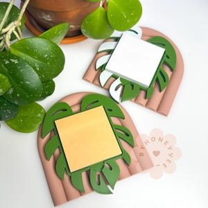 May include: Two decorative monstera leaf-shaped memo pad holders. Each holder has a light brown base with green leaf accents. One holds a stack of white notepaper, the other holds a stack of yellow notepaper. A potted plant is in the background.