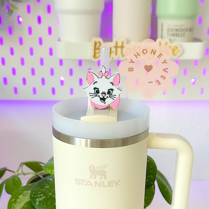 May include: A white and pink cat cartoon character straw topper on a white and cream Stanley travel mug with a black handle. The mug has the Stanley logo on the front.