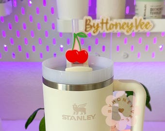 May include: A white Stanley travel mug with a black logo and a cherry-shaped straw topper. The mug has a handle and a white lid. The straw topper is red and green.