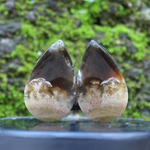 May include: A pair of teardrop-shaped agate earrings with brown, white, and pink patterns. The earrings are made of natural stone and have a polished finish.