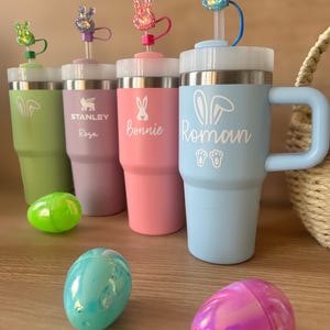 Puede incluir: Una colección de vasos aislados de colores pastel con adornos en forma de conejito y pajitas. Cada vaso está personalizado con un nombre o logotipo. Huevos de Pascua en colores a juego están esparcidos frente a los vasos. Los vasos tienen un asa y un borde de acero inoxidable.