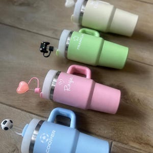 May include: Four insulated tumblers with handles and straws, each featuring a unique topper and personalized name. The colors are light blue with a soccer ball, pink with a heart, light green with a star and Franklin, and cream with a heart.