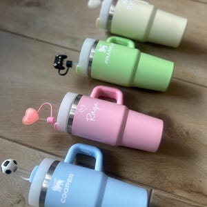 Pode incluir: Quatro canecas isoladas em cores pastel: verde claro, rosa e azul claro. Cada caneca tem uma alça, uma borda prateada e uma tampa branca. As canecas são personalizadas com nomes e um logotipo. Uma tem um topo de palha em forma de coração.