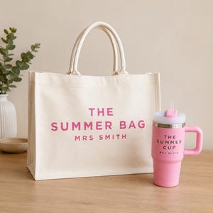 May include: A cream-coloured canvas tote bag with two handles, featuring the pink text THE SUMMER BAG and MRS SMITH. Beside it is a pink stainless steel tumbler with a handle, a clear straw, and the text THE SUMMER CUP and MRS SMITH.