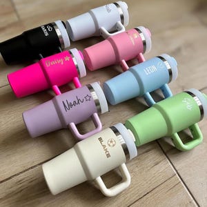 Pode incluir: Uma variedade de canecas isoladas em várias cores, incluindo preto, branco, rosa, azul claro, roxo e verde. Cada caneca tem uma alça e uma borda prateada. Algumas canecas são personalizadas com nomes como "Noah", "Daisy" e "Blake".