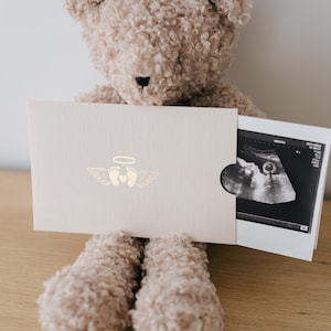 May include: A light beige card holder featuring a gold foil graphic of baby feet with wings and a halo, partially revealing a black and white ultrasound scan inside, held by a textured light brown teddy bear with a dark nose and eyes.