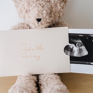 May include: A light beige card with the text "Just a little note to say..." printed in a cursive font, featuring a cutout that reveals a black and white ultrasound scan, positioned in front of a fuzzy, light brown teddy bear with a black nose.